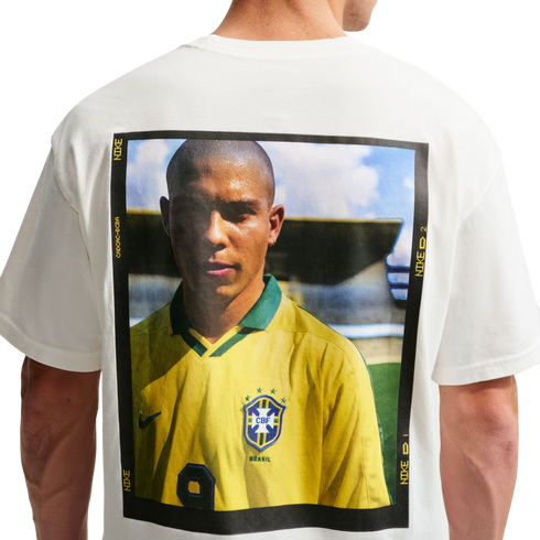 The back of a white t-shirt featuring a large, rectangular graphic print of Brazilian soccer legend Ronaldo Nazário. He is wearing a yellow Brazil national team jersey, and the portrait is enclosed in a black frame with yellow Nike branding text.