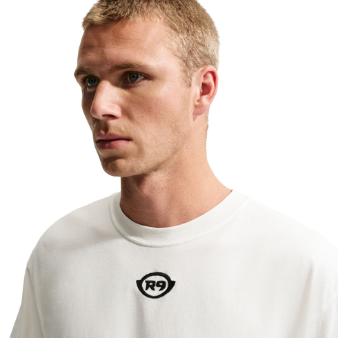 A medium shot of a man with short blonde hair wearing a white t-shirt with a small black "R9" logo in the center, set against a solid black background.