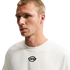 A medium shot of a man with short blonde hair wearing a white t-shirt with a small black "R9" logo in the center, set against a solid black background.