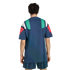 Rear view of a person wearing a navy blue t-shirt with green, white, and red geometric panels on the shoulders and matching blue striped shorts.