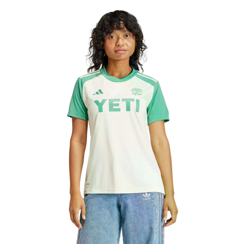 A person wears a white and green soccer jersey with a "YETI" logo and blue jeans against a black background.
