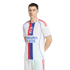 A man models the white Olympique Lyonnais home soccer jersey featuring a central red and blue stripe, team crest, and Emirates sponsor. He stands against a solid black background.