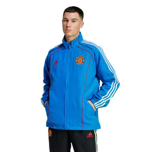 A man models a blue Adidas Manchester United track jacket and black track pants against a plain white background. The jacket features white stripes and red accents.