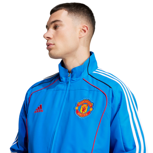 A man wearing a blue Manchester United track jacket with white stripes and a team crest, set against a plain white background.
