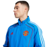 A man wearing a blue Manchester United track jacket with white stripes and a team crest, set against a plain white background.