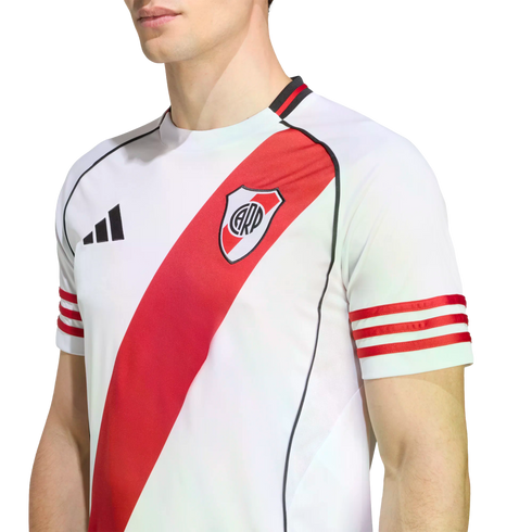 A person wears a white River Plate football jersey featuring a red diagonal sash, three red stripes on the sleeves, and the Adidas logo. Set against a plain white background.