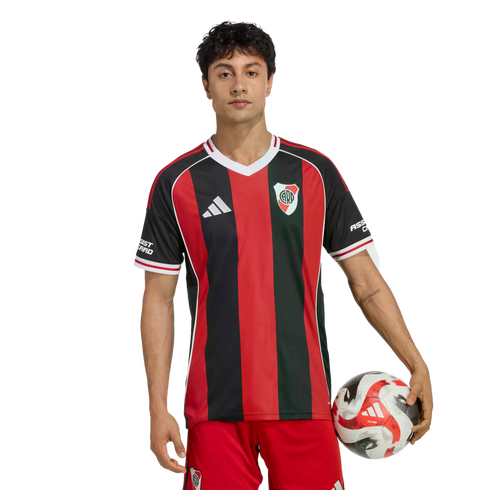 A man in a red and black striped River Plate soccer jersey and red shorts holds a soccer ball against a white background.