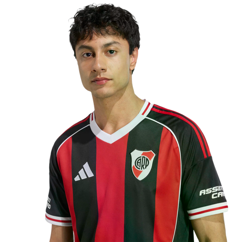 Portrait of a young man with dark wavy hair wearing a red and black striped River Plate soccer jersey against a plain white background.