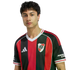 Portrait of a young man with dark wavy hair wearing a red and black striped River Plate soccer jersey against a plain white background.