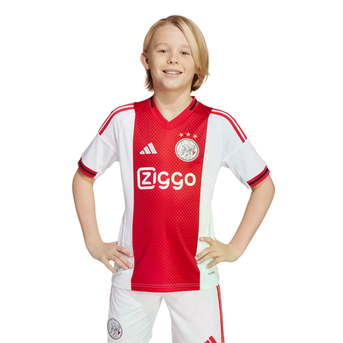 A smiling blond boy in a red and white Ajax home football kit stands with hands on his hips against a plain white background.