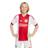 A smiling blond boy in a red and white Ajax home football kit stands with hands on his hips against a plain white background.