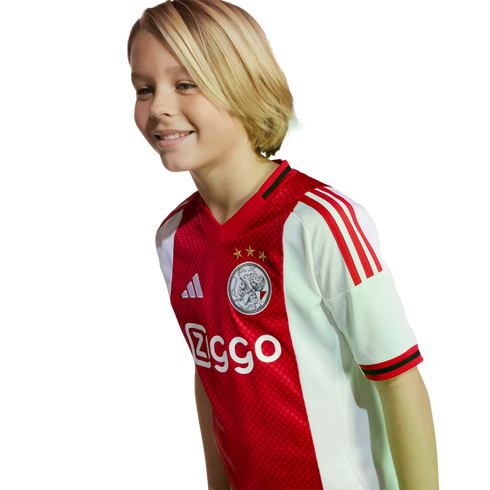 A smiling blonde child wearing a red and white Ajax football jersey.