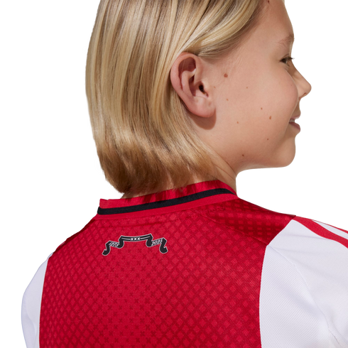 A blonde child from behind wearing a red and white soccer jersey. A small black banner on the collar reads "1275 XXX 2025".