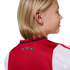 A blonde child from behind wearing a red and white soccer jersey. A small black banner on the collar reads "1275 XXX 2025".
