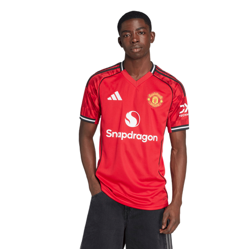 A Black man stands against a white background wearing a red Manchester United home jersey with the Snapdragon logo and black pants.