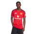 A Black man stands against a white background wearing a red Manchester United home jersey with the Snapdragon logo and black pants.