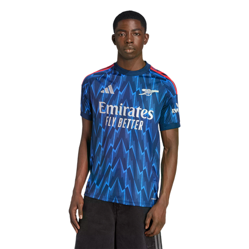 Man modeling a blue Arsenal football jersey with a zigzag pattern and white Emirates logo on a black background.