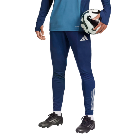 A person in blue athletic wear and black soccer cleats holds a soccer ball under their arm against a solid black background.