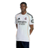 A young Black man in a white Real Madrid soccer jersey with orange accents and sponsor logos, standing with hands behind his back against a white background.