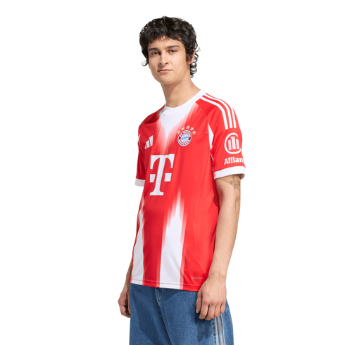A young man with dark curly hair stands against a white background, wearing a red and white Bayern Munich soccer jersey and blue jeans.