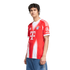 A young man with dark curly hair stands against a white background, wearing a red and white Bayern Munich soccer jersey and blue jeans.