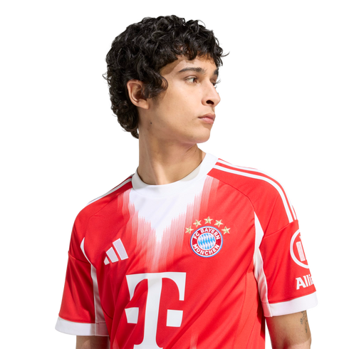 A young man with dark curly hair wears a red and white FC Bayern Munich soccer jersey. He looks off-camera against a plain white background.