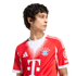 A young man with dark curly hair wears a red and white FC Bayern Munich soccer jersey. He looks off-camera against a plain white background.