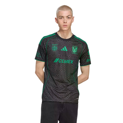 A young man with short blonde hair wearing a black and green Tigres UANL soccer jersey and black jeans, standing against a black background.