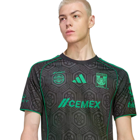 A person with short blonde hair wearing a grey and green patterned Tigres UANL soccer jersey against a black background.