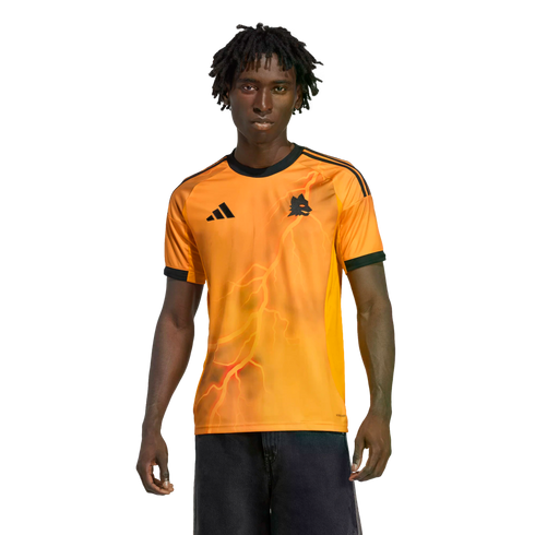 A young man wearing a bright orange Adidas jersey with a lightning bolt graphic and wolf crest, standing against a solid black background.