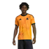 A young man wearing a bright orange Adidas jersey with a lightning bolt graphic and wolf crest, standing against a solid black background.