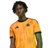 A man with dreadlocks wearing an orange Adidas jersey with a wolf crest and lightning bolt graphics against a black background.