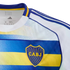 Close-up of a white, blue, and yellow Boca Juniors jersey featuring the CABJ crest on a blue band, three blue stripes on the shoulder, and internal Adidas branding.
