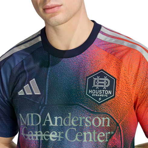 A person wears a Houston Dynamo FC soccer jersey with a pixelated blue-to-orange gradient, featuring the team crest, Adidas logo, and MD Anderson Cancer Center sponsorship.