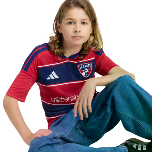 A young person with long hair sits in an FC Dallas jersey and baggy blue jeans against a black background.