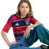 A young person with long hair sits in an FC Dallas jersey and baggy blue jeans against a black background.