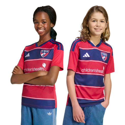 Two smiling young fans wearing red and blue striped FC Dallas home jerseys and blue shorts, posing against a black background.