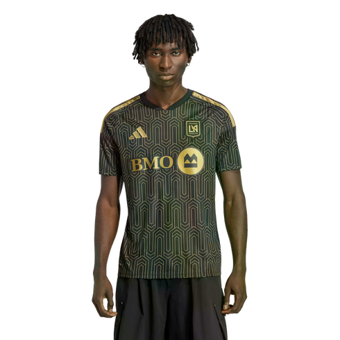 A man with dreadlocks wears a black and gold LAFC soccer jersey with a geometric pattern against a solid black background.