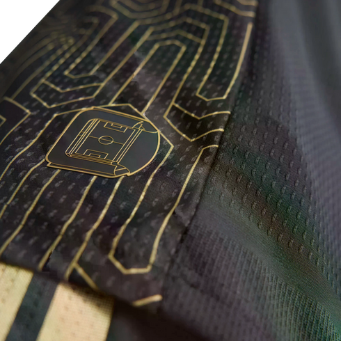 Close-up of a black sports jersey with gold geometric line patterns and a small badge of a football pitch. The fabric has a fine mesh texture.
