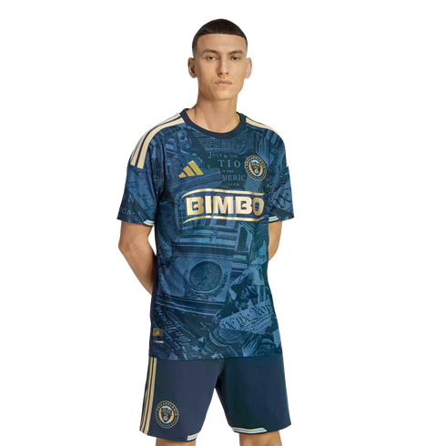 A man models a navy blue and gold Philadelphia Union soccer kit featuring historic Philadelphia-themed graphics, standing against a black background.