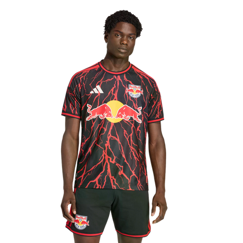 A man models a black Red Bull New York soccer jersey with a red lightning bolt pattern and matching black shorts against a black background.