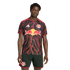 A man models a black Red Bull New York soccer jersey with a red lightning bolt pattern and matching black shorts against a black background.