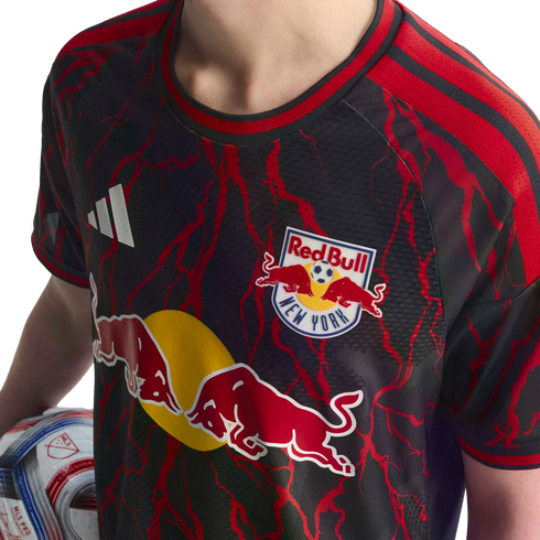 A person wears a black New York Red Bulls soccer jersey with red lightning patterns and prominent team logos, while holding an MLS soccer ball.