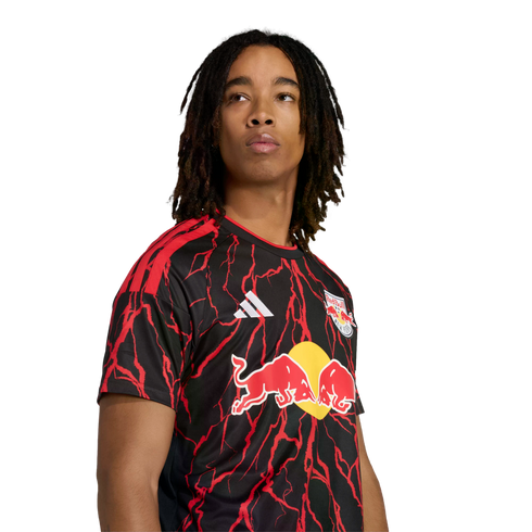 A person with long dreadlocks wearing a black and red lightning-patterned New York Red Bulls soccer jersey against a black background.