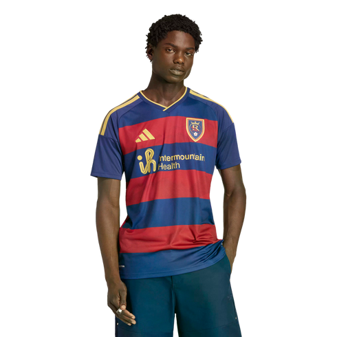 A young man models a Real Salt Lake soccer jersey with red and blue stripes and yellow accents against a black background.