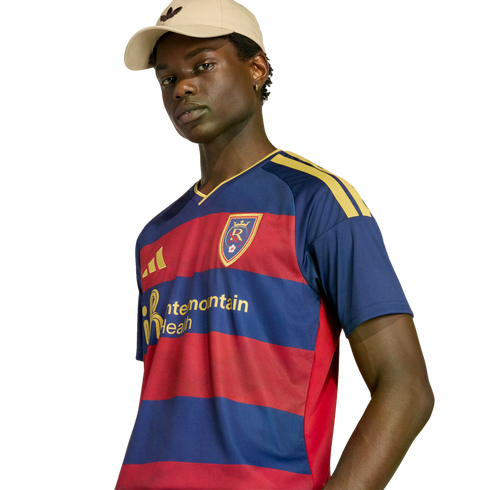 A man in a beige cap and a navy and red striped Real Salt Lake soccer jersey with yellow accents, posed against a black background.