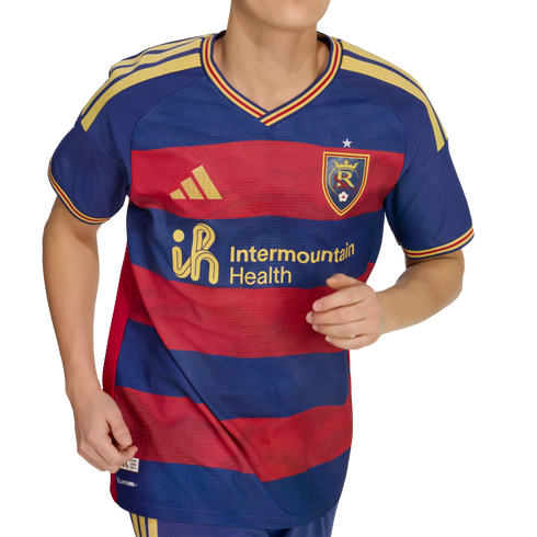 A person wears a blue and red striped soccer jersey featuring gold Adidas and Intermountain Health logos, with yellow shoulder stripes and a team crest.