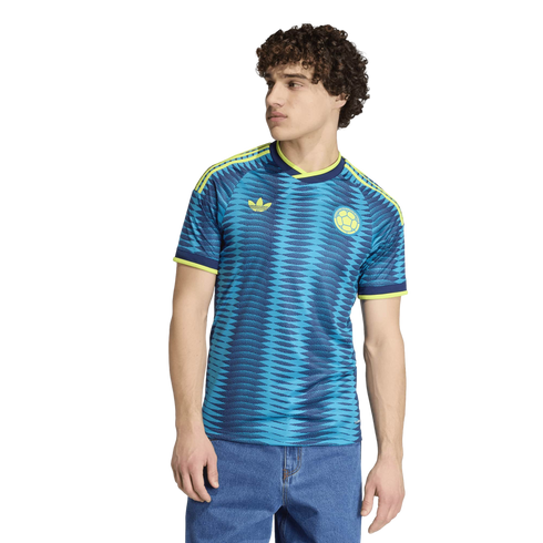 Young man with curly hair wearing a blue patterned soccer jersey and blue jeans, standing against a black background.