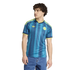 Young man with curly hair wearing a blue patterned soccer jersey and blue jeans, standing against a black background.