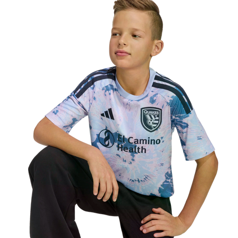 A young boy with brown hair kneeling, wearing a tie-dye San Jose Earthquakes soccer jersey and black pants against a black background.